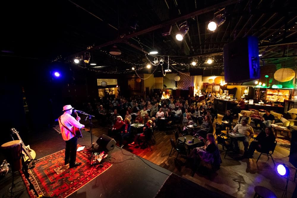 A person wearing a hat, white longsleeve shirt and black pants performs to a seated crowd in a large venue.  A person wearing a hat, white longsleeve shirt and black pants performs to a seated crowd in a large venue.