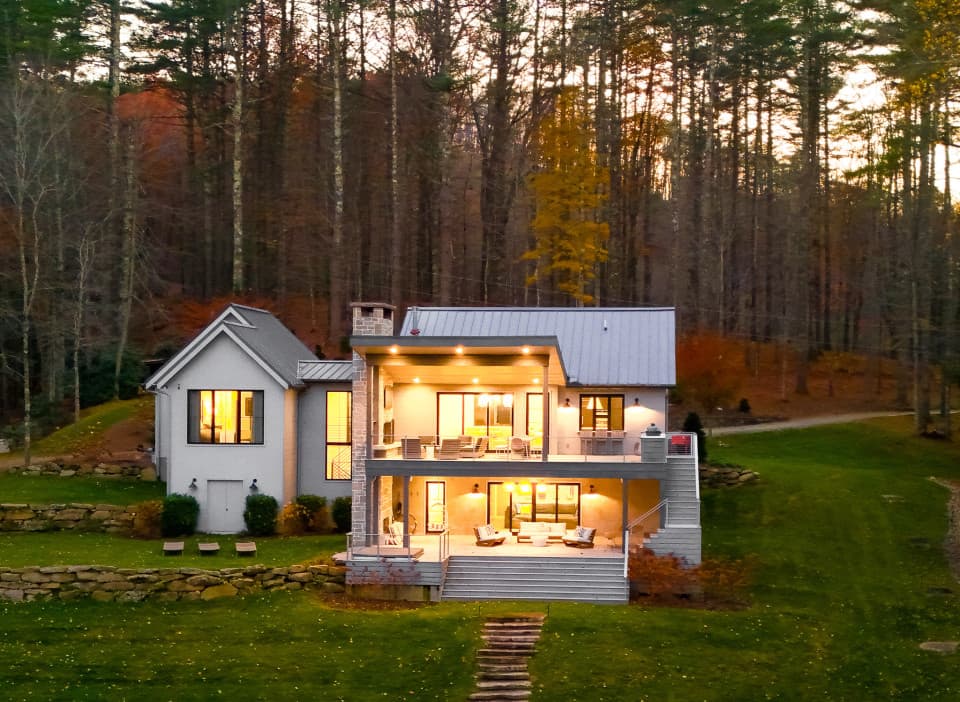 Modern luxury mountain home surrounded by tall trees at dusk, featuring expansive covered decks, warm interior lighting, stone retaining walls, and a peaceful forest setting in the Blue Ridge Mountains. Modern luxury mountain home surrounded by tall trees at dusk, featuring expansive covered decks, warm interior lighting, stone retaining walls, and a peaceful forest setting in the Blue Ridge Mountains.