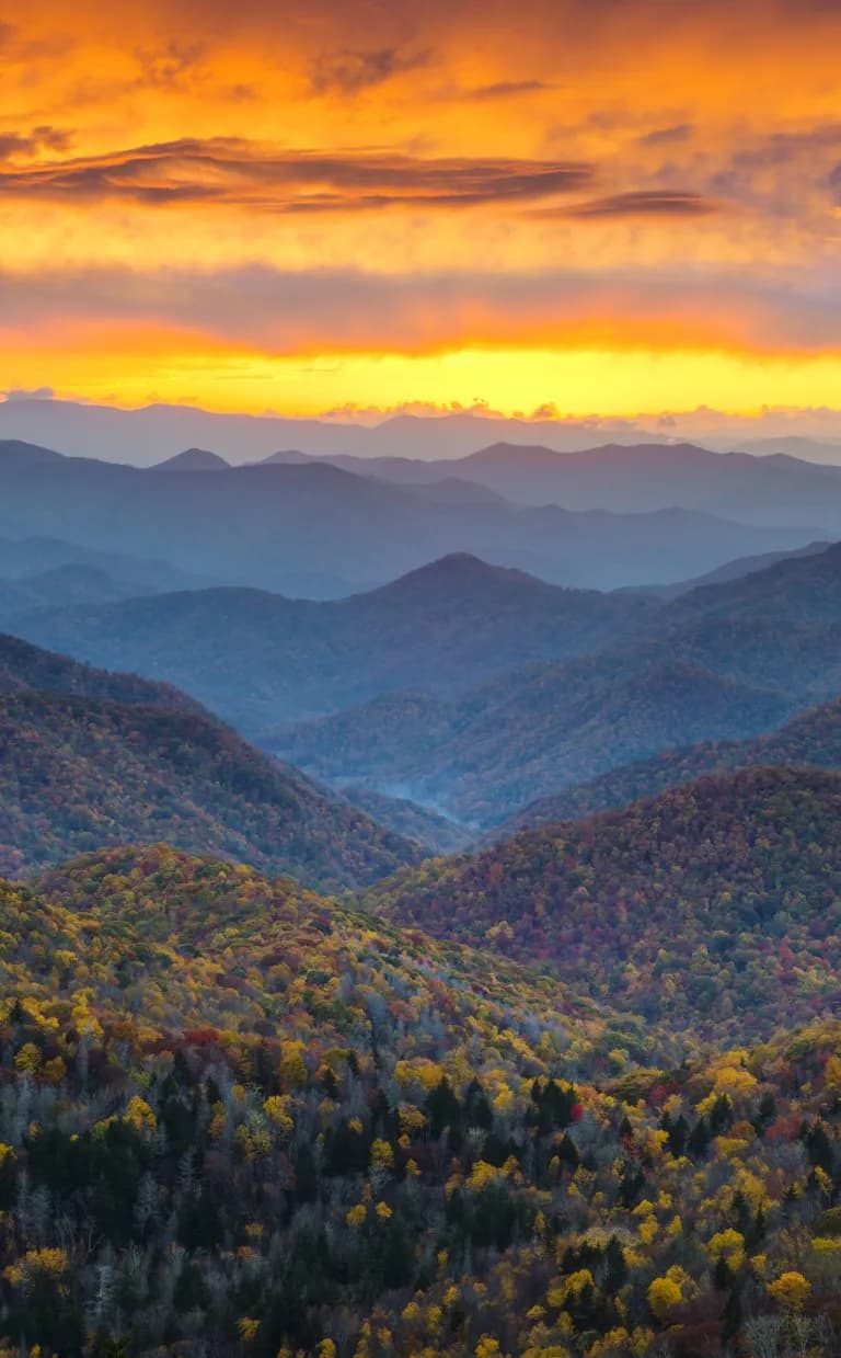 Faces of The History of Great Smoky Mountains National Park Blue mountains below a sun-streaked orange sky in Asheville